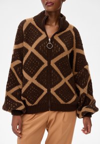 Kobieta ubrana w brązowy i jasnobrązowy sweter na zamek w romby z balonowymi rękawami oraz jasnobrązowe spodnie.