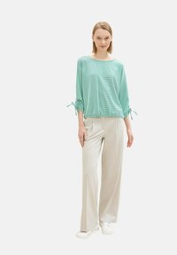 Helles türkisfarbiges, strukturiertes Blusenshirt mit weitem Ausschnitt und gerafften Ärmeln, die an den Bündchen gebunden werden, kombiniert mit hellbeigen, weit geschnittenen Hosen und weißen Schuhen.