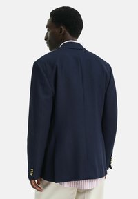 Blazer blu navale realizzato in tessuto testurizzato, con un'unica fessura, due tasche laterali e bottoni dorati sui polsini. Vista posteriore.