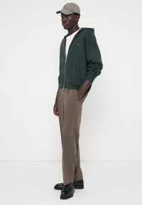 Felpa con zip verde scuro e tasche a canguro, abbinata a pantaloni marrone chiaro. Un cappellino beige con un motivo sottile completa l'outfit.