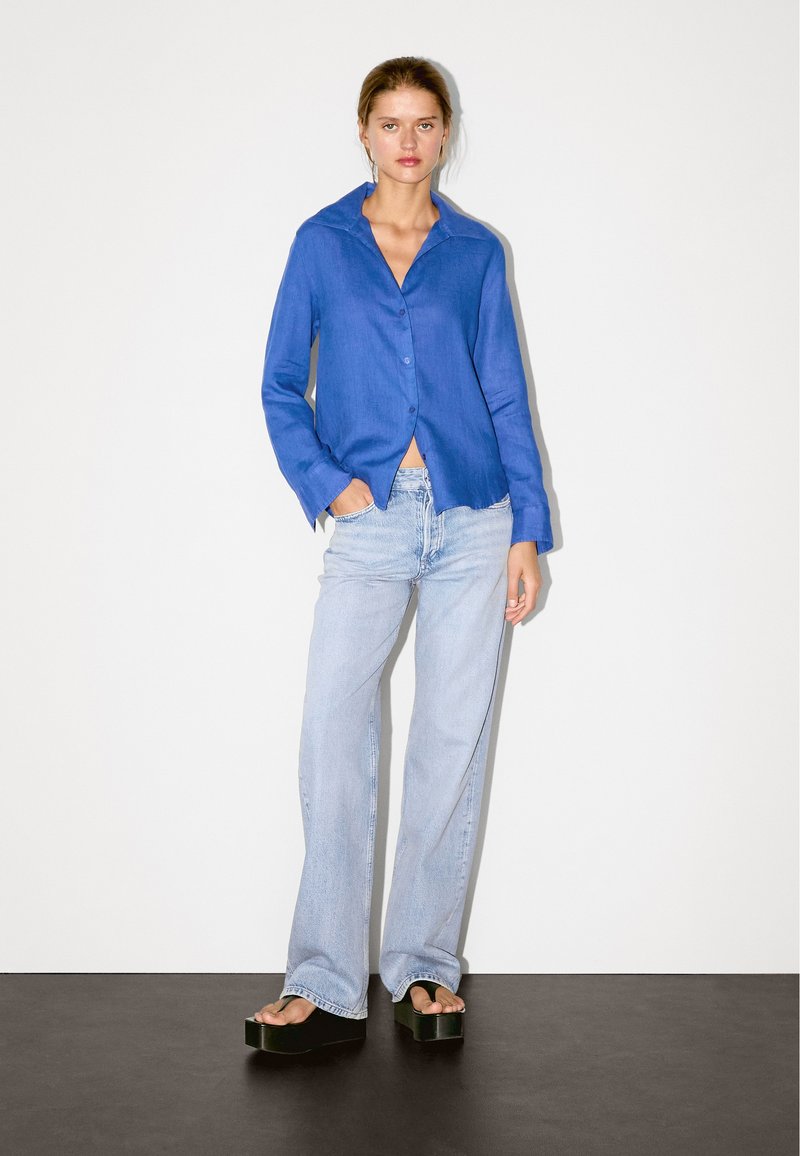 Blaues Hemd mit Kragen, kombiniert mit hellblauen weit geschnittenen Jeans und schwarzen Plateausandalen, vor einem neutralen Hintergrund.