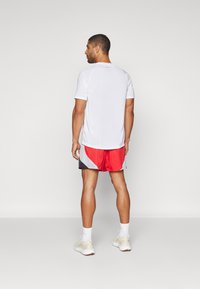 Hvit kortermet treningsskjorte, rød og svart shorts med sidesømdetaljer, hvite joggesko med lette såler, synlig ribbet tekstur på skjorten.