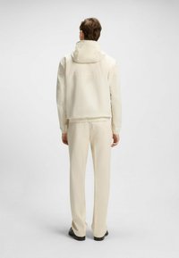 Giacca cropped di un beige chiaro con cappuccio, polsini elastici e una texture liscia; abbinata a pantaloni affusolati nello stesso tono.