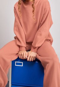 Felpa rosa oversize e pantaloni abbinati, realizzati in tessuto morbido, con spalle scese e maniche ampie, poggiati su una scatola blu.