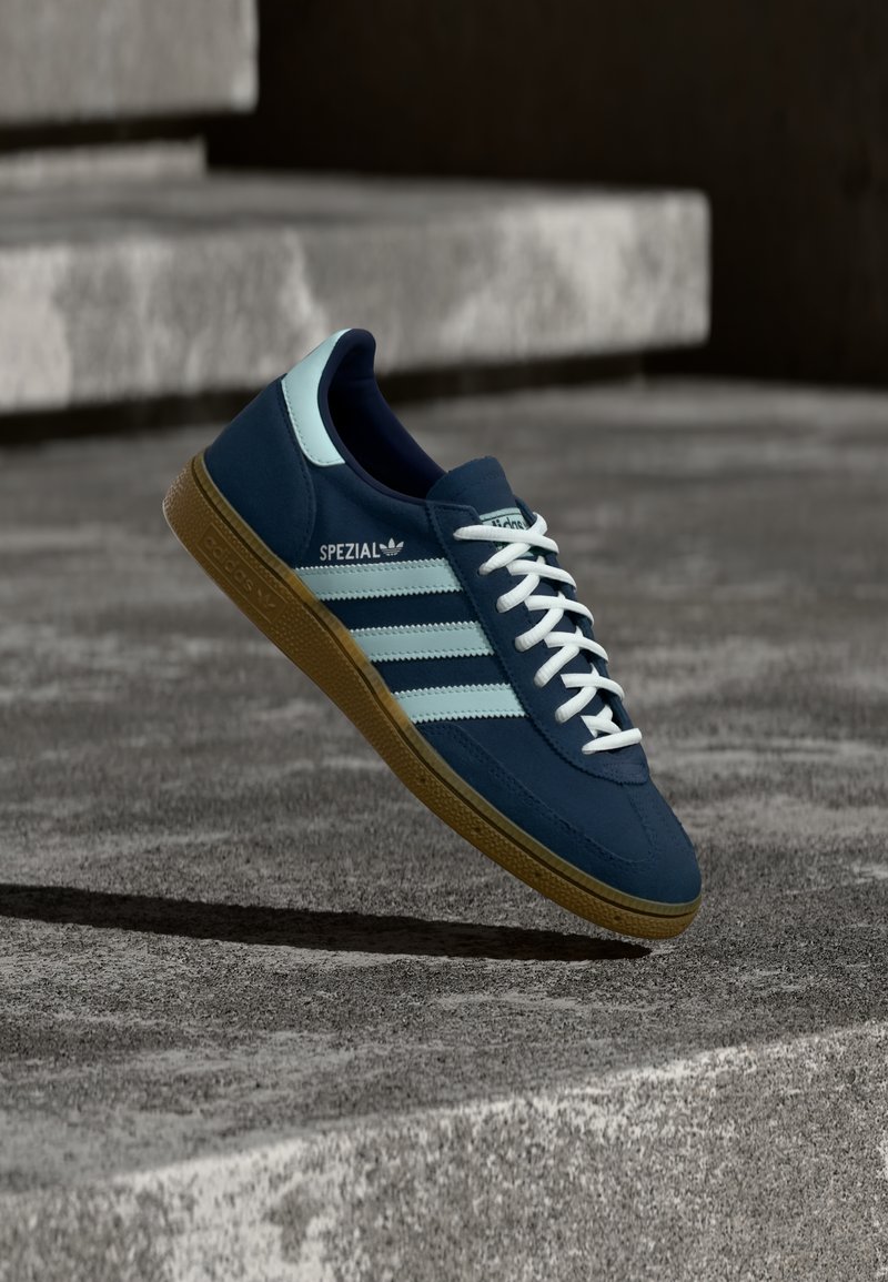 Baskets Adidas Spezial bleues avec des rayures et des lacets blancs, semelle en gomme, flottant au-dessus d'une surface en béton rugueuse et des marches en arrière-plan.
