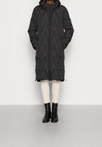 Longue doudoune noire avec capuche, dotée d'une fermeture éclair à l'avant, de coutures diagonales et de poches latérales. Portée avec des leggings crème et des bottes noires.