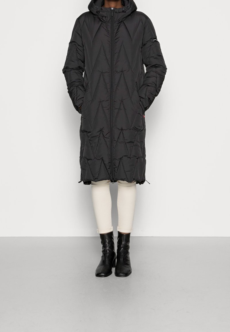 Longue doudoune noire avec capuche, dotée d'une fermeture éclair à l'avant, de coutures diagonales et de poches latérales. Portée avec des leggings crème et des bottes noires.