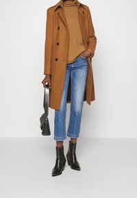 Brązowy płaszcz z mieszanki wełny w podwójnej bretońskiej konstrukcji, w połączeniu z jasnobrązowym swetrem, niebieskimi krótkimi dżinsami i czarnymi botkami na obcasie.
