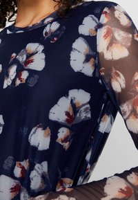 Blus med blommönster i marinblått och långa ärmar i genomskinligt mesh. Har en rund halsringning och mjukt, texturerat tyg med kontrasterande detaljer.