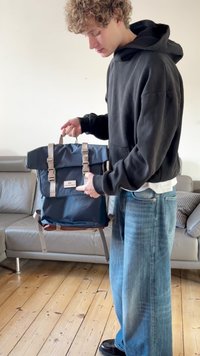 Mochila azul con cierre enrollable, correas beige, hebillas y parche con logo en el frente. Fabricada con material resistente, presenta una forma estructurada y un diseño práctico.