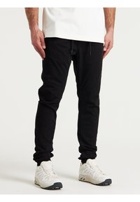 Černé joggingové kalhoty s elastickými manžetami, tkaničkovým pasem a švy, spárované s bílými sportovními botami s tmavými podrážkami.