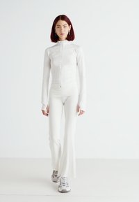 Giacca sportiva bianca con zip e pantaloni flare abbinati. Il materiale appare liscio ed elasticizzato. Sneakers scure visibili. Sfondo semplice.