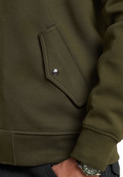 Sweatshirt verde-oliva com tecido texturizado, apresentando um bolso em forma de diamante com fecho de pressão e um design de punho canelado.