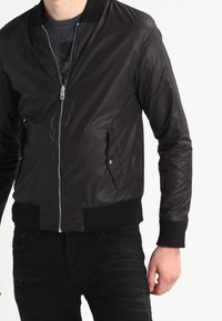 Svart bomberjacka med dragkedjestängning, ribbade ärmslut och nederkant samt sidofickor. Materialen verkar släta och något glänsande.