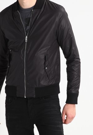 Zwarte bomberjack met een ritssluiting aan de voorkant, ribbelboorden en -zoom, en zijzakken. Materialen lijken glad en licht glanzend.