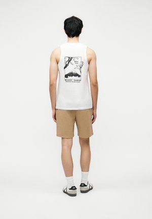 Ung mann sett bakfra iført hvit ermeløs skjorte med svart grafikk av Mount Fuji, beige shorts, hvite sokker og joggesko.