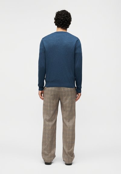 Άνδρας με σγουρά μαλλιά που φοράει μπλε πουλόβερ με μακριά μανίκια και μπεζ καρό παντελόνι, στέκεται έχοντας την πλάτη προς το μέρος μας σε απλό φόντο.