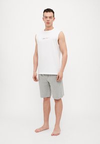 Hvit ermeløs bomullsbluse med "Calvin Klein"-logo og grå bomullsshorts. Modellen står barbeint på en ensfarget bakgrunn.