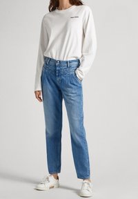 Person som bär en vit långärmad skjorta med "Pepe Jeans"-logotyp, högmidjade blåjeans och vita sneakers, står med ena handen i fickan.