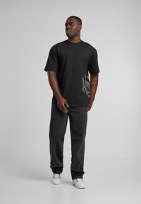Black cotton t-shirt with short sleeves, featuring a distinctive gray graphic design on the lower side, paired with dark gray pants and white sneakers.