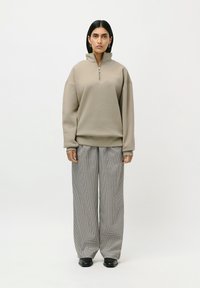 Kvinne står iført en beige quarter-zip genser, svart og hvit rutete vide bukser og svarte ankelstøvler på hvit bakgrunn.