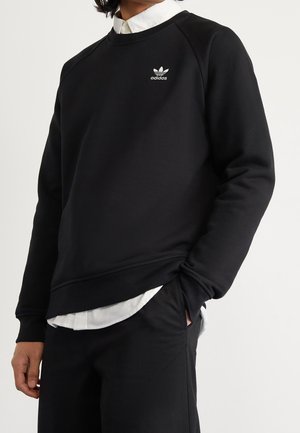 Person iført sort Adidas sweatshirt over hvid skjorte med hænder i sorte bukselommer, beskåret ved skuldre og lår.