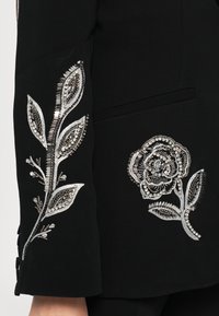 Blazer in tessuto nero con decorazioni floreali argentate ricamate sulle maniche, caratterizzato da perline e paillettes, che esaltano i dettagli e la texture.