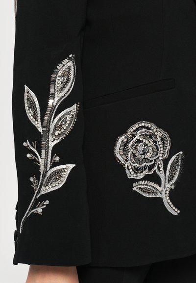 Black fabric blazer with embellished silver floral designs on the sleeves, featuring bead work and sequins, accentuating detail and texture.