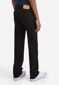 Persona vistiendo jeans negros de pierna recta con bolsillos traseros y zapatillas Converse All Star blancas, de pie contra un fondo simple.