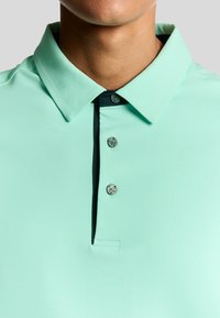 Mintgrön polo t-shirt med klassisk krage, tre knappar och en kontrasterande marinblå placket med emblemdetaljer. Slät textur.