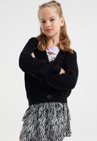 Cardigan nero in maglia con chiusura a bottoni, dotato di maniche ampie. Abbinato a una maglietta grigia e a una gonna a strati con stampa zebra bianco e nero.