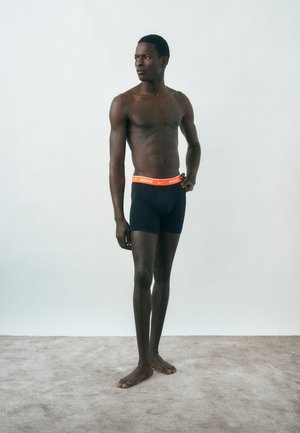 Άνδρας χωρίς πουκάμισο, όρθιος ξυπόλητος σε χαλί, φοράει μαύρα εσώρουχα Nike με πορτοκαλί ζώνη, κοιτάζοντας προς τα αριστερά σε απλό φόντο.