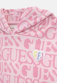 Rózsaszín kapucnis pulóver, amelyen a fehér "GUESS" logó mintázata ismétlődik. Kiemelkedő szív alakú logó díszítéssel, színes betűzéssel az elején.