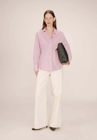 Camicia a righe rosa con maniche lunghe, abbinata a pantaloni bianchi a gamba larga. Tiene una clutch in pelle nera. Sfondo semplice.