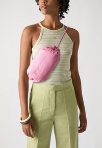 Rosa skinnveske med snøring, båret på siden. Modellen har på seg et stripete tanktop og lysegrønne, høytlivsbukser.