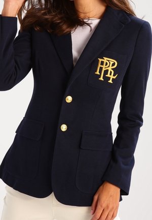 Vrouw draagt een marineblauwe blazer met gouden knopen en een goud geborduurd logo op het borstzakje, gecombineerd met een wit shirt en lichte broek.