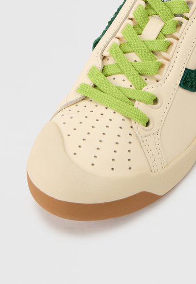 Εκρού μύτη παπουτσιού με τρύπες, ανοιχτοπράσινα κορδόνια, σκουροπράσινη υφασμάτινη λεπτομέρεια και καφετί σόλα ορατή σε λευκό φόντο.