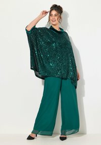 Smaragdgrøn sequin poncho med en blød velourkrave, kombineret med flydende bredbundede bukser i en matchende nuance og sorte hæle.