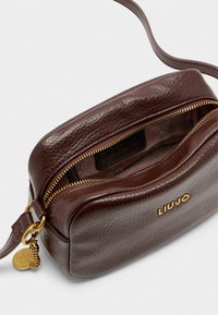 Smeđi kožni crossbody ranac za leđa s zaobljenim oblikom, zlatnim zatvaračem i utisnutim unutrašnjim dijelom. Sadrži zlatni logo i ukrasnu hardveru.