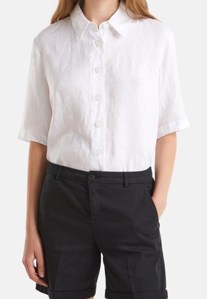 Kobieta ubrana w białą koszulę na krótki rękaw z guzikami, włożoną do czarnych, dopasowanych spodenek, z rękami w kieszeniach, na jednolitym tle.