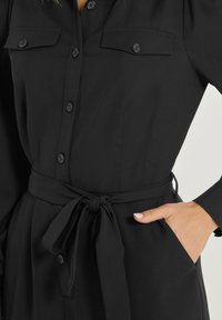 Vestido negro de manga larga con cinturón anudado en la cintura, que presenta dos bolsillos en el pecho y cierre de botones en la parte delantera. Tejido suave y texturizado.