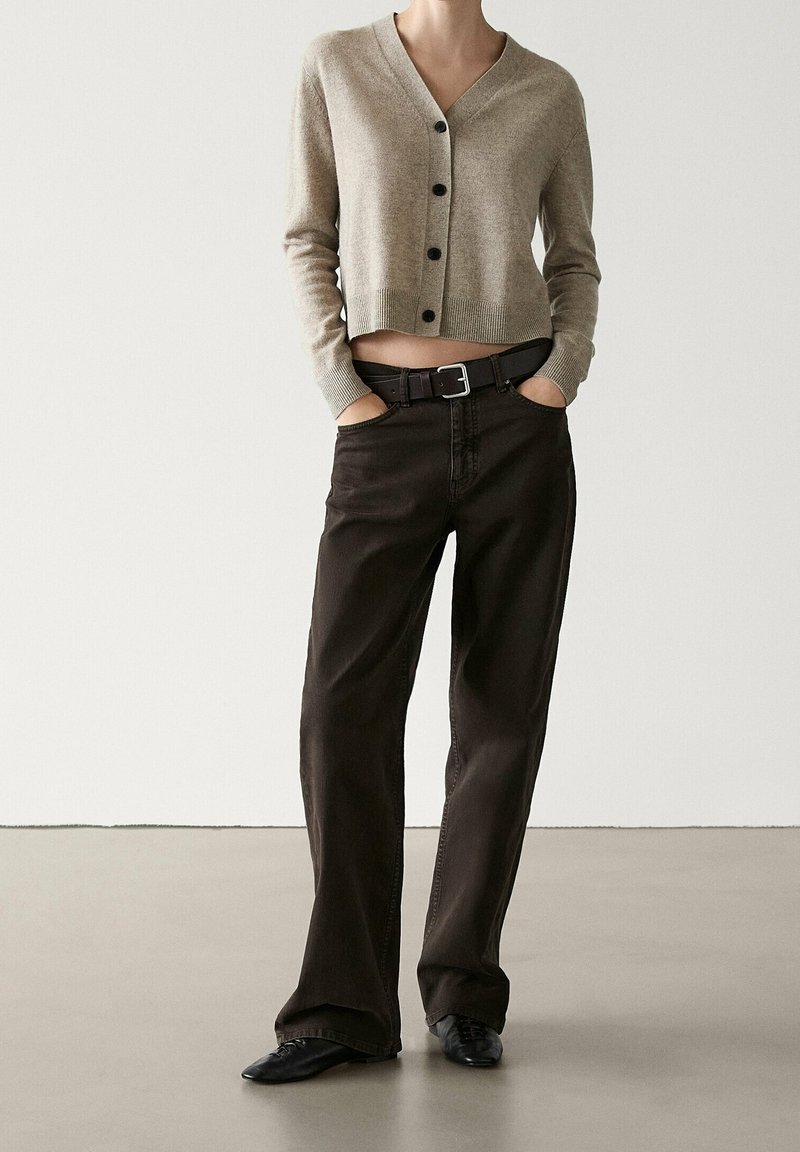 Cardigan beige court avec boutons noirs, associé à un pantalon large marron foncé et une ceinture noire. Texture lisse et coupe décontractée.