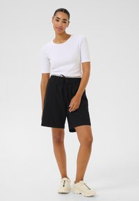 Shorts noirs avec une taille élastique et deux poches avant, associés à un t-shirt blanc ajusté. Le modèle porte des baskets de sport crème.