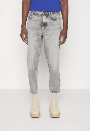 Mand iført lysegrå falmede jeans, hvide ankelstøvler og en blå skjorte, stående mod en ensfarvet hvid baggrund.