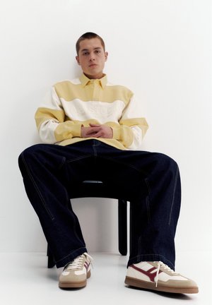 Junger Mann sitzt auf einem schwarzen Stuhl mit weit auseinander gespreizten Beinen, trägt ein gelb-weiß gestreiftes Hemd, dunkle Jeans und beige Sneakers mit roten Akzenten.