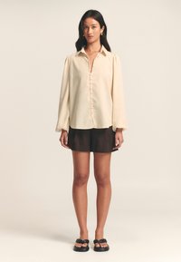 Cream blouse with a collar, button-down front, and puff sleeves, paired with dark shorts and black sandals. Smooth texture, minimal design.