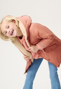 Doudoune rose avec une capuche en fausse fourrure, déboutonnée pour révéler un hoodie rose superposé. Un jean ajusté bleu clair complète la tenue.