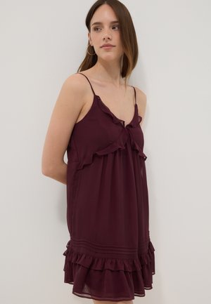 Jeune femme en robe sans manches violette foncée à volants avec fines bretelles, debout devant un fond clair uni, regardant sur le côté.