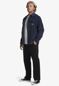 Navy corduroy shirt jacket with two chest pockets and logo detail, worn over a light t-shirt, paired with black pants and sneakers.