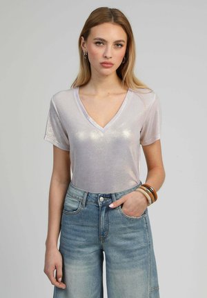 Femme aux longs cheveux blonds portant un haut argenté brillant à manches courtes et col en V, un jean taille haute bleu et des bracelets en bois, debout avec une main dans la poche.